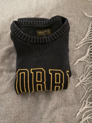 Mörkblå stickad tröja Morris - Snygg mörkblå stickad tröja från Morris med gul broderad text 'MORRIS SINCE 1968' på bröstet. Tröjan har rund halsringning och ribbade muddar vid ärmslut och nederkant. Perfekt för en avslappnad men stilren look. Passar M och en tajtare L.