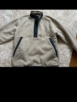 Beige fleece tröja med svarta detaljer - Mysig beige fleece tröja med svarta detaljer vid krage, knappslå och fickor. Tröjan har hög krage, knappstängning framtill och två snedställda fickor. Perfekt för kalla dagar och enkel att matcha med jeans eller mjukisbyxor.