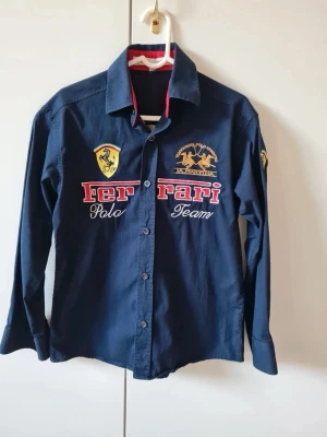 Barn skjorta med Ferrari Polo Team tema - Marinblå långärmade skjorta från samarbete mellan La Martina och Ferrarri Polo Team Skjorta har broderade logotyper. Storlek 146/152.