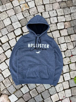 Blå Hollister hoodie med tryck - Snygg blå hoodie från Hollister med vit och svart text på bröstet där det står 'Hollister California' samt logga. Hoodien har en stor ficka framtill och justerbar huva med svarta snören. Perfekt för en avslappnad och cool stil.