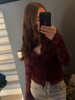 Vinröd spetsblus med volang - Säljer en vinröd blus i spets med volangkant längs knäppningen. Blusen har lång ärm och är lätt genomskinlig, vilket ger en trendig och elegant look. Perfekt att styla med jeans eller kjol för en cool outfit.