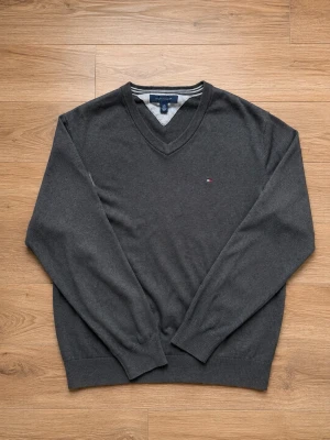 Mörkgrå V-ringad tröja Tommy Hilfiger - Klassisk mörkgrå V-ringad tröja från Tommy Hilfiger med diskret logga på bröstet. Tröjan har långa ärmar, ribbade muddar och är tillverkad i mjukt material som passar perfekt till lager-på-lager-stil.