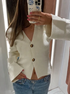 Gina tricot cardigan  - Stilren krämvit kofta med djup V-ringning och två stora bruna knappar framtill. Modellen har vida ärmar och lätt utsvängd passform nedtill. Perfekt att styla med jeans för en trendig look.