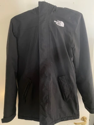 Svart parkas från The North Face - Svart parkasjacka från The North Face med huva och fuskpälsdetalj. Jackan har två snedställda fickor framtill och logga på både bröst och rygg. Klassisk rak modell med dragkedja och vindslå, perfekt för kyliga dagar.
