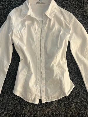 Vit figursydd skjorta med knappar - Säljer en vit figursydd skjorta med lång ärm och knappar framtill. Skjortan har en figurnära passform och en snygg krage. Perfekt till både jeans och kostymbyxor. Materialet känns som bomull och ger en fräsch look.