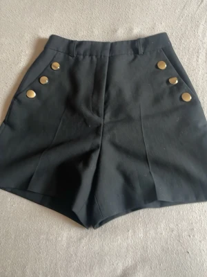 Svarta shorts med guldknappar - Stilrena svarta shorts med hög midja och dekorativa guldknappar framtill. Klassisk design med bälteshällor och en ficka bak. Perfekta för dig som vill ha en chic och enkel look.