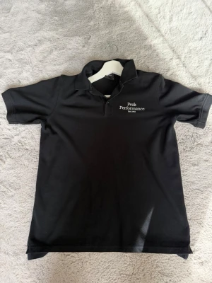 Svart Peak Performance pikétröja - Klassisk svart pikétröja från Peak Performance med vit logga på bröstet. Tröjan har krage och korta ärmar, perfekt för en clean och sportig look. Tillverkad i mjukt material som känns skönt mot huden.