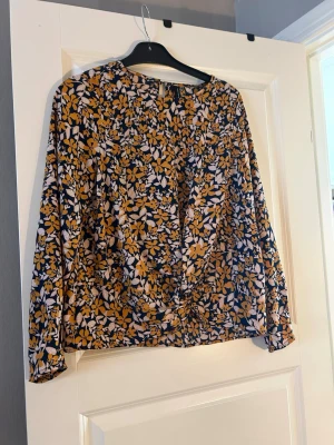 Blommig blus med knutdetalj - Snygg blus med blommigt mönster i senapsgult, vitt och marinblått. Blusen har lång ärm och en trendig knutdetalj framtill. Tillverkad i ett lätt och luftigt tyg som ger en skön känsla. Perfekt för dig som gillar färg och mönster.