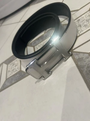 Svart bälte med stor silverspänne - Snyggt svart bälte med ett stort, blankt silverspänne i form av ett H. Bältet har en stilren och modern look som passar till många outfits. Perfekt accessoar för att lyfta din stil.