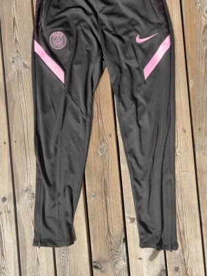 Svarta PSG träningsbyxor med rosa detaljer - Snygga svarta träningsbyxor från Nike med PSG-logga på ena benet och rosa detaljer längs sidorna. Byxorna har smal passform, resår i midjan och är tillverkade i ett lätt och stretchigt material som passar perfekt till träning.