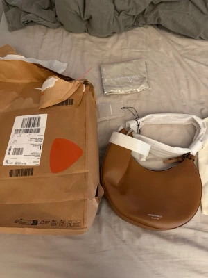 Brun axelremsväska Emporio Armani - Snygg halvmåneformad handväska från Emporio Armani i slätt brunt läder. Väskan har silverfärgad dragkedja och justerbar axelrem. Passar perfekt för en present till en tjej eller nån tjejkompis 