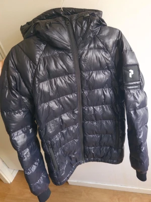 Peak performance jacka - Snygg svart pufferjacka med glansig finish och huva. Jackan har dragkedja framtill, ribbade muddar vid ärmsluten och en ficka med dragkedja på ena ärmen. Perfekt för kalla vinterdagar och ger en modern look.