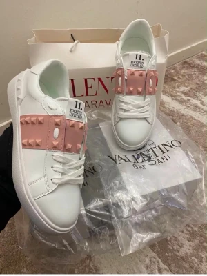 Vita sneakers med rosa nitar Valentino - Snygga vita sneakers från Valentino med rosa band och coola nitar över ovandelen. Skorna har en chunky sula med rutmönster undertill och vita skosnören. Tillverkade i slätt läder för en clean look och detaljer som sticker ut.