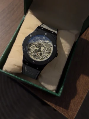 Klocka - Stilren svart klocka med skeletturverk som visar de inre mekanismerna. Klockan har rund boett, silverfärgade detaljer och svart armband. Perfekt accessoar för dig som gillar modern design och vill sticka ut.