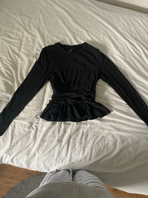 Svart volangtopp från Gina Tricot - Svart långärmad topp från Gina Tricot med volang nertill och figurnära passform. Tillverkad i mjuk bomull, perfekt för en stilren och trendig look. Rund halsringning och enkel design som passar till mycket.