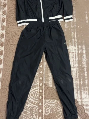 Svart träningsset från Lacoste - Lacoste traksuit som är använd bara några gånger, inget är skadad bara att på vänsta och högra knäna så har färgen gott bort lite men det syns inte mycker när man har på sig den, båda är i storlek M och priset kan sänkas och skickar dem samma dag vid köp.