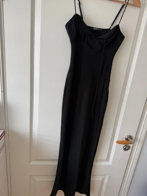Svart långklänning från Gina Tricot - Svart långklänning från Gina Tricot med smala axelband och snygg snörning vid bysten. Klänningen har en enkel, elegant siluett och är tillverkad i ett mjukt, följsamt material som känns skönt mot huden. Perfekt för dig som gillar minimalistisk stil. Jag har använt denna klänning 1 gång och den böev tyvärr för liten. 