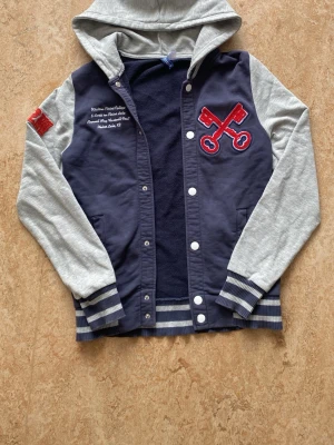 Blå och grå hoodie med tryck - Snygg hoodie i collegemodell med marinblå kropp och ljusgrå ärmar och huva. Har broderat rött emblem med nycklar på bröstet och vit text på ena sidan. Ribbstickade muddar och nederkant med ränder. Perfekt för en sportig och avslappnad stil. Storlek S