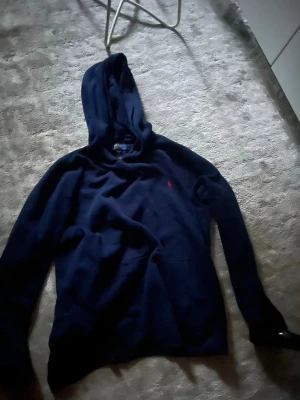 Mörkblå hoodie från Polo Ralph Lauren - Snygg mörkblå hoodie från Polo Ralph Lauren med klassisk huva och den ikoniska röda loggan broderad på bröstet. Tillverkad i mjukt material som känns skönt mot huden. Perfekt för en avslappnad och stilren look.