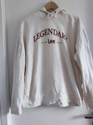Vit hoodie från Lee med tryck - Snygg vit hoodie från Lee med texten 'LEGENDARY' och 'Lee Denim' i vinrött framtill. Klassisk huva med dragsko och regular fit. Tillverkad i mjukt bomullsmaterial, perfekt för en avslappnad och trendig look.