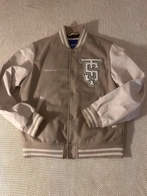Beige och vit bomberjacka - Snygg beige och vit bomberjacka med broderad detalj på bröstet och randiga muddar. Jackan har tryckknappar framtill, två fickor och kontrasterande vita ärmar. Perfekt för en trendig och avslappnad stil. 
