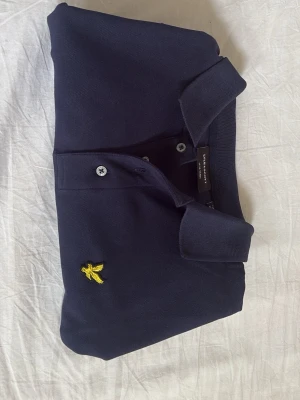 Marinblå pikétröja från Lyle & Scott - Snygg marinblå pikétröja från Lyle & Scott med klassisk krage och två knappar framtill. Tröjan har det ikoniska gula fågelmärket broderat på bröstet. Tillverkad i mjuk bomull och har korta ärmar för en clean och stilren look.