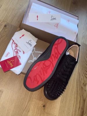 Christian Louboutin sneakers - Säljer ett par svarta sneakers från Christian Louboutin med ikonisk röd sula och svarta nitar på tån. Skorna har en klassisk låg siluett och är tillverkade i mocka med svarta skosnören. Perfekta för dig som vill sticka ut med exklusiv design.