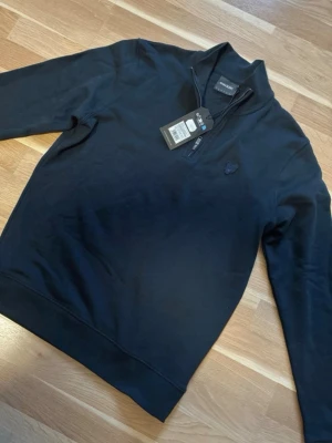 Marinblå half zip från Lyle & Scott - Snygg marinblå tröja från Lyle & Scott med half zip och diskret broderad logga på bröstet. Tillverkad i mjuk bomullsblandning och har ribbade muddar vid ärmslut och nederkant. Perfekt för en clean och stilren look.