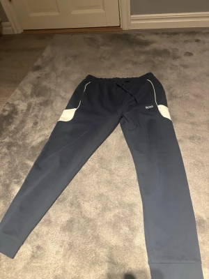 Marinblå mjukisbyxor från BOSS - Snygga marinblå mjukisbyxor från BOSS med vita detaljer vid fickorna och logga på vänster sida. Byxorna har dragsko i midjan och muddar vid bensluten. Perfekta för chill dagar eller när du vill ha en sportig look.