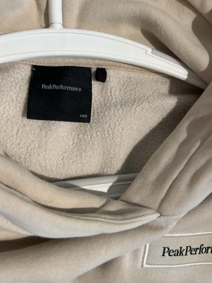 Beige hoodie från Peak Performance - Snygg beige hoodie från Peak Performance med broderad logga på bröstet. Hoodien har en stor huva och är fodrad med mjukt, fluffigt material på insidan för extra komfort. Ribbstickade muddar vid ärmslut och nederkant. Perfekt för chill dagar.storlek 160. Köpt från intersport för 899kr. Tröjan är använd 2 gång och har små fläckar som är enkla att tvätta bort.
