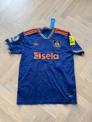 Blå Newcastle matchtröja Elanga 20 - Snygg blå matchtröja från Newcastle United med Elanga och nummer 20 på ryggen. Tröjan har orange detaljer, klubbmärke på bröstet, Premier League-märke på ärmen och sponsorlogga framtill. Tillverkad i lätt och ventilerande material, perfekt för fotboll eller träning. Stolek XL men sitter som L!