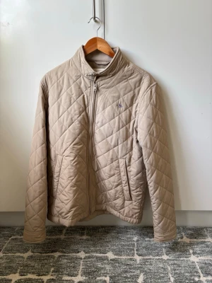 Beige quiltad jacka från Gant - Snygg beige quiltad jacka från Gant med dragkedja framtill och ståkrage. Jackan har två sidofickor och diskret logga på bröstet. Perfekt för dig som gillar stilren och klassisk design.
