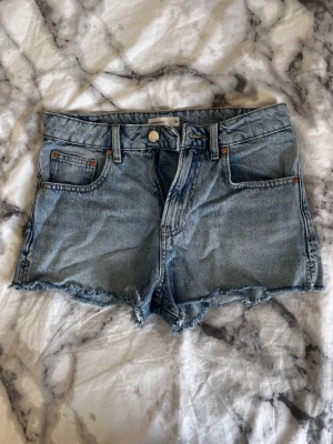 Blå jeansshorts med rå kant - Säljer ett par klassiska blå jeansshorts med rå, fransig kant och femficksmodell. Perfekta för varma dagar och enkel att matcha med allt.