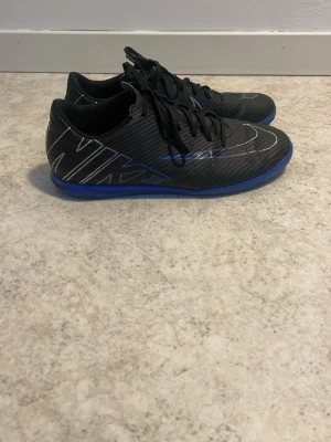 Svarta Nike inomhusfotbollsskor - Säljer ett par svarta Nike inomhusfotbollsskor med blå sula och en Nike-text samt Nike-logga på sidorna i färgen silver/blå. Perfekt för till exempel futsal eller bara användas som gympaskor.