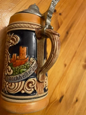 Tysk ölsejdel med lock och motiv - Dekorativ tysk ölsejdel i keramik med detaljerat handtag och metallock. Utsmyckad med färgglada motiv av människor och ett slott samt texten 'nach heißer Jagd'. Perfekt som samlarobjekt eller inredningsdetalj.