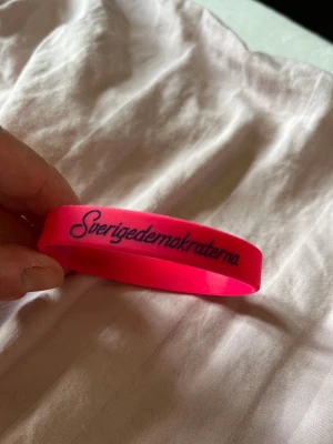 Rosa silikonarmband ”sverigedemokraterna” - Coolt rosa armband i silikon med svart texten 'Sverigedemokraterna' tryckt på utsidan. Armbandet är flexibelt och har en enkel, rund form som passar de flesta handleder. Perfekt för dig som vill sticka ut med en färgstark accessoar.