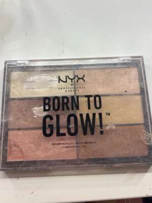 NYX Born To Glow Highlighterpalett - Säljer en NYX Born To Glow highlighterpalett med sex skimrande nyanser i guld, champagne, brons och rosé. Perfekt för att skapa glow och highlighta ansiktet. Paletten har en rektangulär plastförpackning med genomskinligt lock.