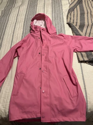 Rosa regnjacka från Cross Sportswear - Snygg rosa regnjacka från Cross Sportswear med huva och tryckknappar framtill. Jackan har två fickor och är tillverkad i ett vattenavvisande material med vit insida. Perfekt för regniga dagar när du vill hålla dig torr och se cool ut.