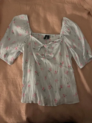 Blommig blus - Supersöt blus med små rosa blommor. Puffärmar och fyrkantig ringning. Knyt framme vid bysten och lätt rynkat tyg. Resår på ryggen. 