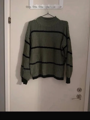Olivgrön stickad tröja med svarta ränder - Säljer en olivgrön stickad tröja med breda svarta horisontella ränder. Tröjan har en hög, ribbad halsringning och långa ärmar med ribbade muddar. Materialet är grovstickat och ger en avslappnad och mysig känsla.