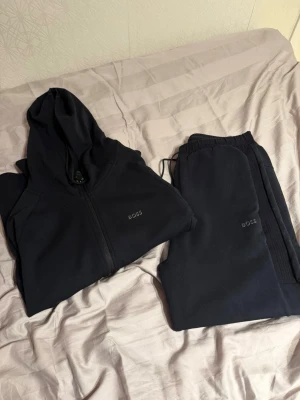 Svart BOSS set med hoodie och byxor - Säljer ett svart set från BOSS med hoodie och matchande byxor. Hoodien har dragkedja och diskret BOSS-logga på bröstet, byxorna har resår i midjan och logga på benet. Mjukt material, perfekt för chill dagar.