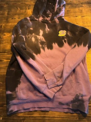 Lila och svart tie-dye hoodie - Säljer en unik hoodie i lila och svart tie-dye med stor färgglad tryck på ryggen och mindre tryck på bröstet och ärmen. Hoodien har huva och magficka, samt coola grafiska detaljer med texten 'TENTACLES' på ryggen. Perfekt för dig som gillar streetwear och statement-plagg.