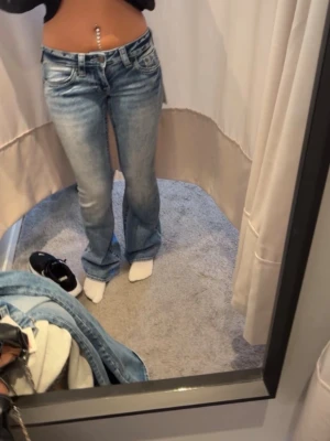Bootcut ljusblå jeans med strass - Säljer ett par ljusblå bootcut jeans med slitningar och snygga strassdetaljer på bakfickorna. Jeansen har låg midja och klassisk femficksmodell. Perfekta för dig som gillar en retro vibe och vill sticka ut lite extra. Ny pris 1399, använda max 2 gånger.