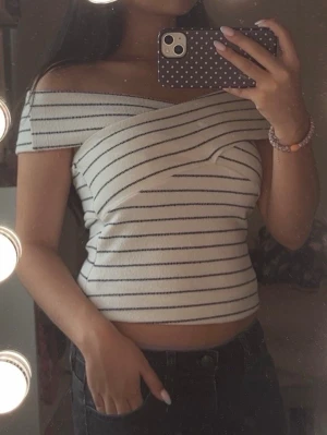 Randig offshoulder topp i vitt och svart  - Använt bara 3 gånger, i princip helt ny! Säljer en snygg offshoulder topp med smala svarta ränder på vit botten. Toppen har en omlott-design över bysten och är croppad. Perfekt för dig som gillar trendiga och unika plagg med en fräsch vibe.