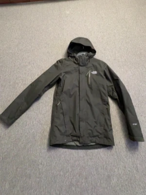 Olivgrön skaljacka från The North Face - Snygg olivgrön regnjacka från The North Face med huva, dragkedja och flera praktiska fickor. Jackan har en minimalistisk design och är tillverkad i ett vattenavvisande material, perfekt för regniga dagar. Logotyp på bröstet och ärmen.
