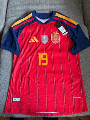 Spaniens landslagströja nr 19 - Röd och marinblå matchtröja från Adidas för Spaniens landslag med gula detaljer och nummer 19. Tröjan har officiella märken, CLIMACOOL-material och Lamine Yamal tryckt på ryggen. Perfekt för fotbollsfans som vill stötta sitt lag.