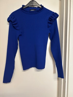 Blå tröja med volang från ONLY - Supersnygg kornblå långärmad tröja från ONLY med volangdetaljer på axlarna och ribbad struktur. Tröjan har en högre hals med liten volangkant och är figurnära i modellen. Perfekt för dig som vill ha något extra till din outfit.