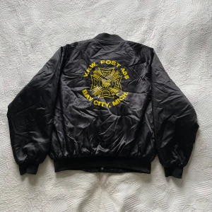 Svart bomberjacka - Retro svart bomberjacka från Nemesis Sportswear med stor V.F.W. POST 485 Bay City, Mich.-logga på ryggen och en liten framtill. Jackan har ribbade muddar, tryckknappar och två fickor framtill. Tillverkad i USA, quiltad insida och cool satin-finish. Säljer då den är för stor för mig. Storlek XL.