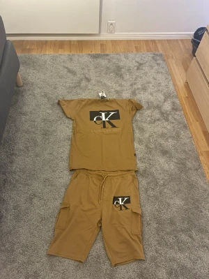 Brunt set från Calvin Klein Jeans - Tvådelat set i brun färg med t-shirt och shorts från Calvin Klein Jeans. Båda plaggen har stor CK-logga i svart och vitt framtill. Shortsen har dragsko i midjan och fickor på sidorna. Perfekt för en avslappnad stil. Prislapparna sitter kvar, aldrig använda.