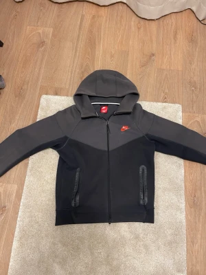 Svart hoodie från Nike med dragkedja - Säljer en svart hoodie från Nike med hel dragkedja framtill och huva. Hoodien har två fickor med dragkedja och en liten röd Nike-logga på bröstet. Perfekt för en sportig och avslappnad stil.